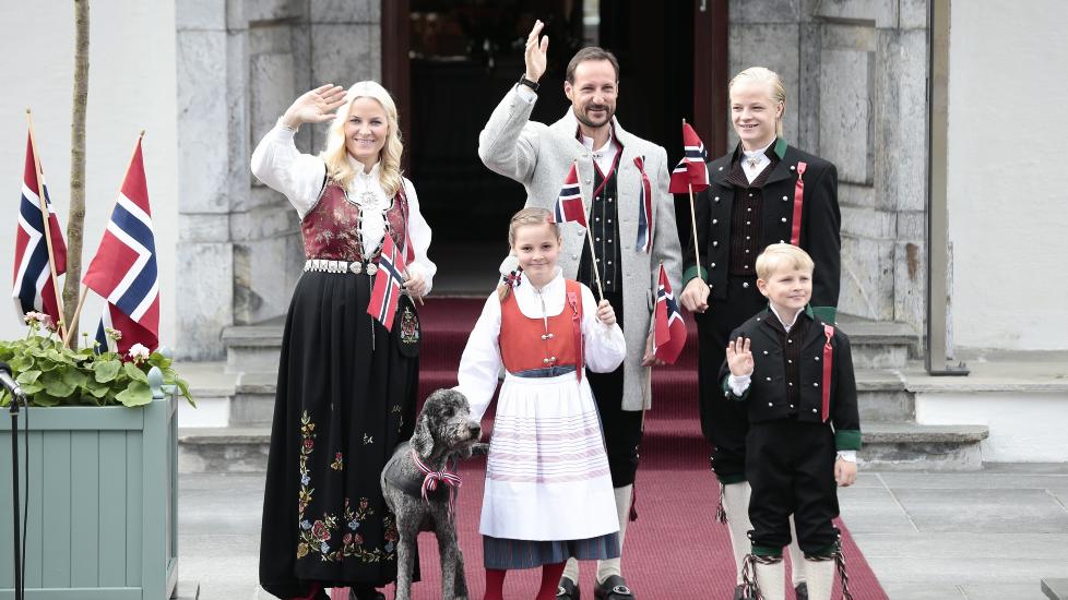 <b>ULIKE BUNADER: </b> 
Kronprins Haakon er kledd i Asker-bunad, kronprinsesse Mette-Marit i Rogalands-bunad, prinsesse Ingrid Alexandra, prins Sverre Magnus, Marius Borg Høiby i bunader fra Vest-Agder. Hunden Milly Kakao hilser sammen med resten av familien på barnetoget utenfor Skaugum for få minutter siden.