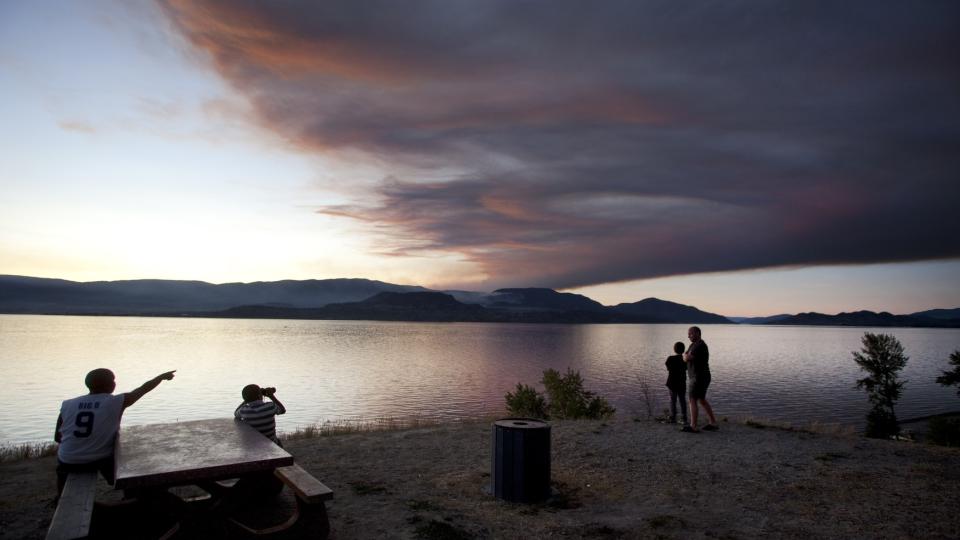 TRE VOLDSOMME BRANNER: Innbyggere i Kelowna by i Canada har samlet seg ved Okanagan-sj�en hvor de kan se r�yken fra skogbrannene stige opp. 10 000 er evakuert som f�lge av brannene og over 6 000 er bedt om � v�re klare til � forlate hjemmene sine. Foto: REUTERS / Andy Clark / SCANPIX